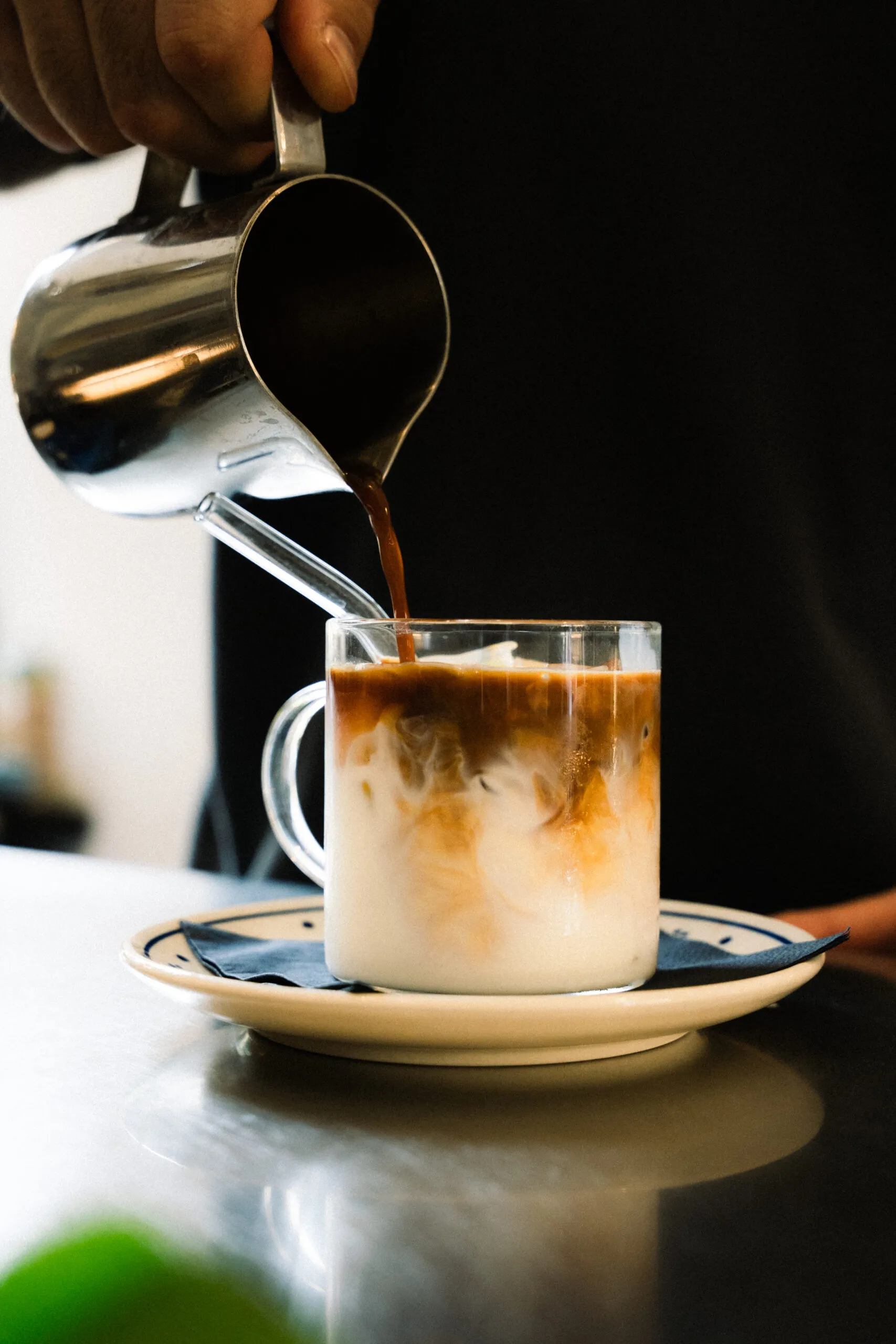 Barista gießt frischen Kaffee aus einem Metallkännchen in ein Glas mit Milch, der sich auf einem Unterteller befindet.